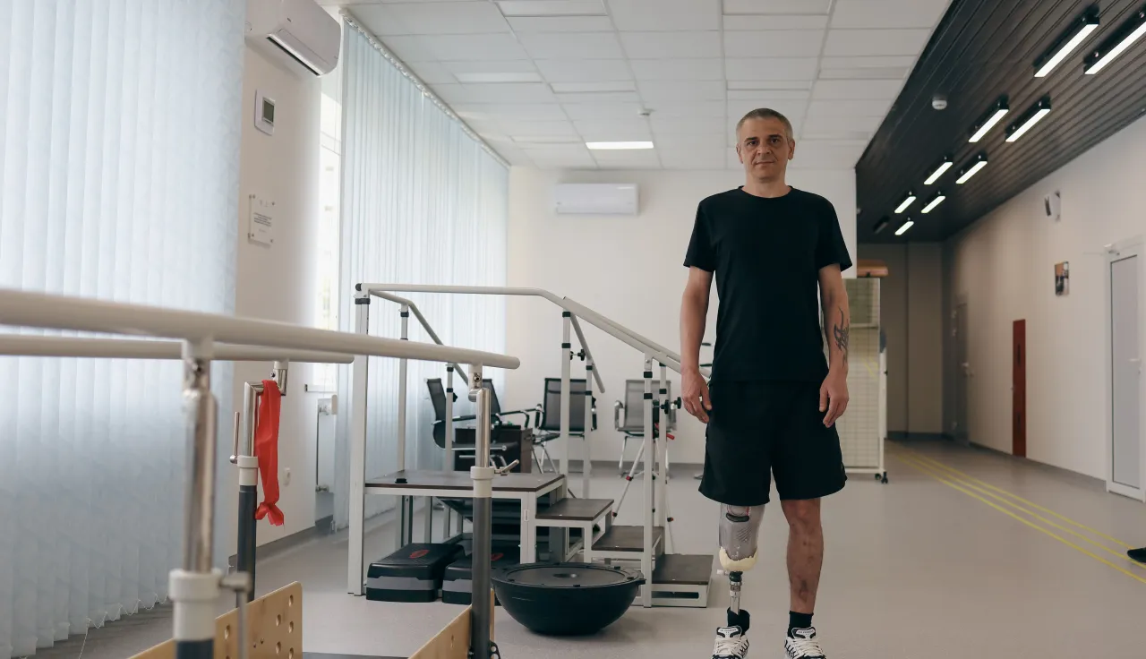 A man with two lower-leg prosthetic limbs stands in a rehabilitation centre beside parallel bars used for gait training. Exercise equipment is visible in the background of the bright therapy room.