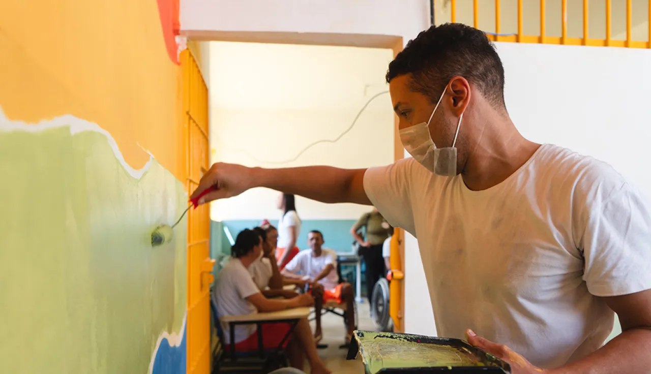 Una persona privada de libertad, con mascarilla, pinta una pared con un rodillo dentro de un centro de detención. En el fondo, otras personas participan en la actividad mientras permanecen en un pasillo del establecimiento.