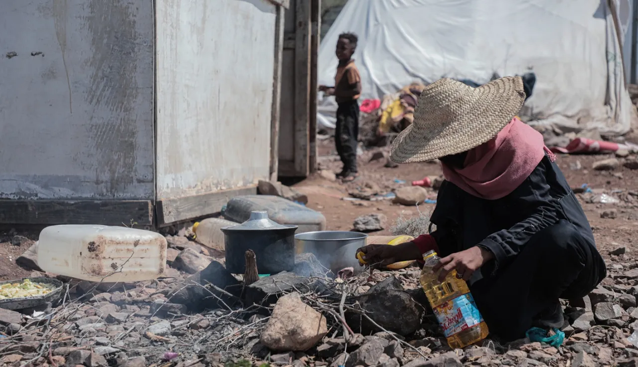 A person cooks over a small outdoor fire among rubble and makeshift shelters.