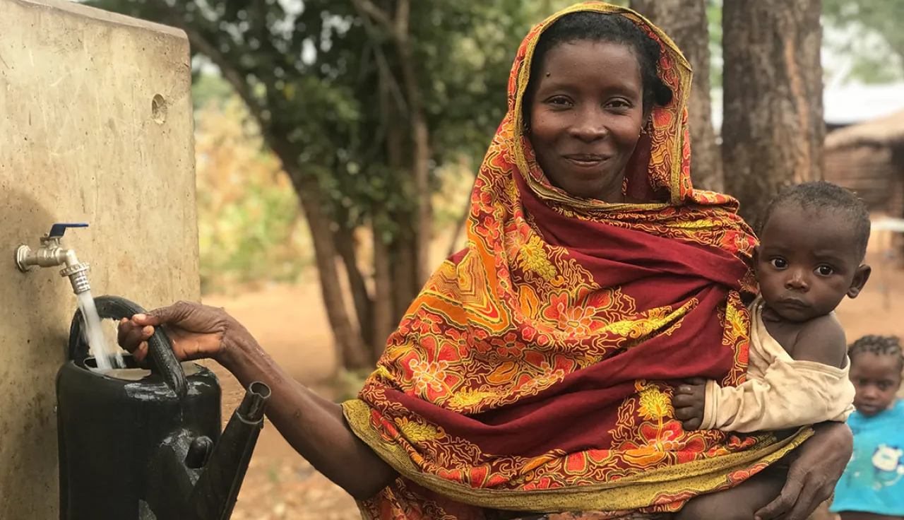 A woman carrying a child fills up a garden sprinkler in her other hand.