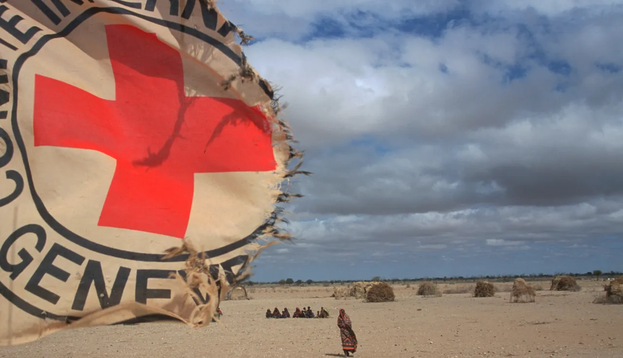 A torn International Committee of the Red Cross (ICRC) emblem hangs in the foreground of a dry, sandy landscape in Ethiopia under a cloudy sky, while a lone person walks in the distance among scattered makeshift shelters.