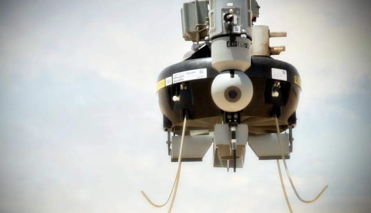 Close-up, low-angle view of a black-and-white mechanical device with a circular central component, metal supports and visible cables, against a pale sky.