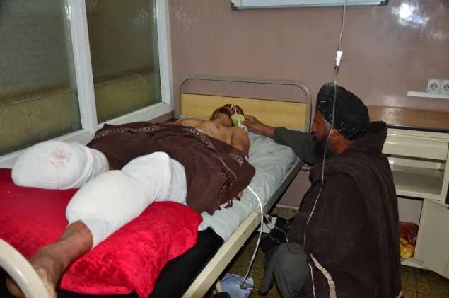 这名年轻的男子因踩上地雷而受伤，右腿截肢，父亲蹲在床前喂他喝果汁，眼神显得很无助。