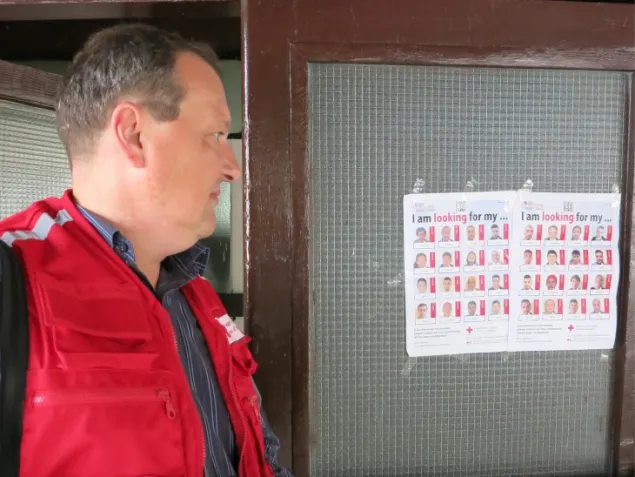 Homem com colete da cruz vermelha olha cartaz com fotos de migrantes.