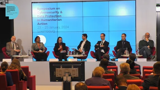Seven panelists sitting on the stage with a blue background and the Symposium's title and date in black text.