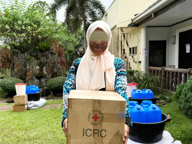 Asniah, smiling, holds an ICRC box, with other donated items in the background.jpg