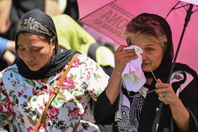 Two women wearing headscarves walk side by side; one holds a pink umbrella while wiping tears with a cloth, and the other looks down with a solemn expression.