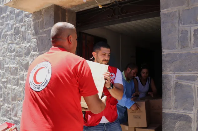 Humanitarian workers wearing Red Crescent and Red Cross vests unload and distribute boxes of aid supplies at a building entrance.