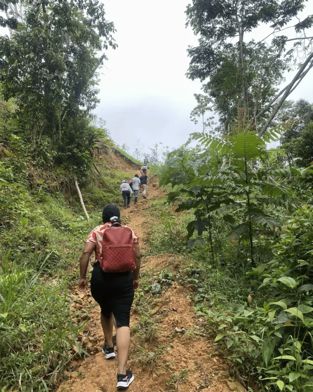 Personas de la comunidad y del CICR recorren a pie un camino rural y montañoso, rodeado de vegetación, en dirección a una zona apartada.
