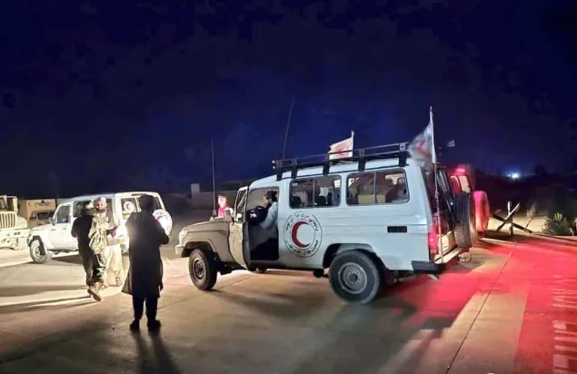 A Red Crescent vehicle is parked on a road at night with its doors open, as humanitarian workers coordinate alongside another vehicle in the background.