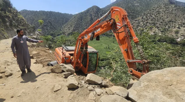 A man wearing a Red Crescent emblem points towards an excavator off a dirt road on a steep hillside in a mountainous area of Afghanistan, following the earthquake.