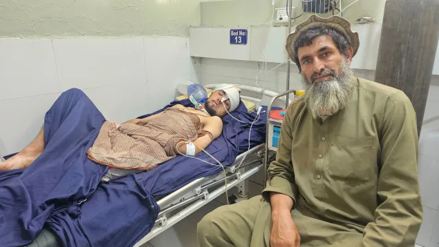 A man sits beside a hospital bed where another man lies injured, covered with a blanket and connected to medical equipment. The patient has a bandage on his head and an oxygen mask, while the man sitting looks at the camera.