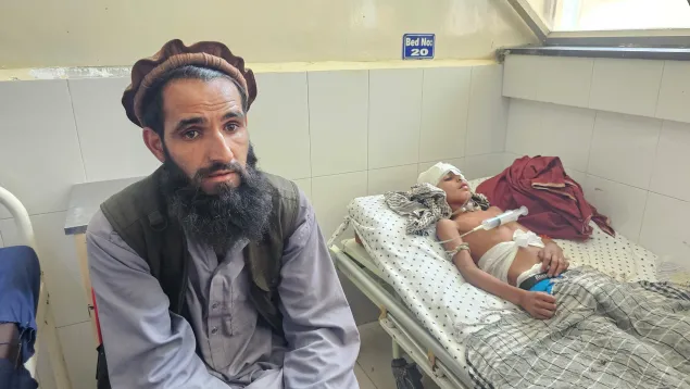 A man sits on a chair beside a hospital bed where a young boy, with bandages on his head and medical tubes attached, lies injured and resting under a blanket.