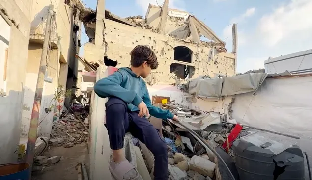 A young boy sits on a concrete ledge amid the rubble of a destroyed building, looking out over the debris and damage around him under a partly cloudy sky.