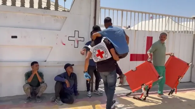 A man carries another person on his back toward a gate marked with a red cross, while others sit or stand nearby, including a man holding two orange stretchers.