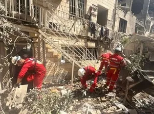 Red Crescent rescue workers in red uniforms clear debris outside a damaged residential building