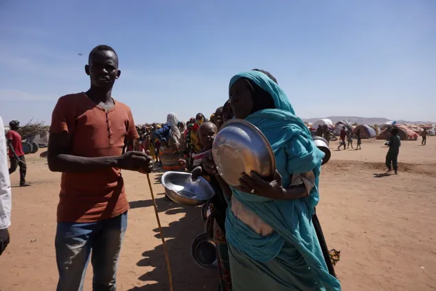 Two people stand in a dusty open area of a displacement site, each holding metal bowls as they wait in a distribution line. Behind them, more people queue with containers, and makeshift shelters stretch across the background under a bright, cloudless sky.