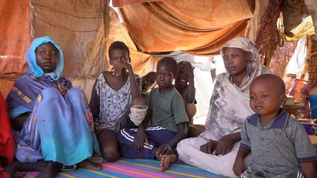 A family sits together inside a makeshift shelter lined with fabric and tarps. Two women and four children sit close on a woven mat, looking toward the camera. The light filters through the patched fabric walls, highlighting the crowded, fragile living conditions.