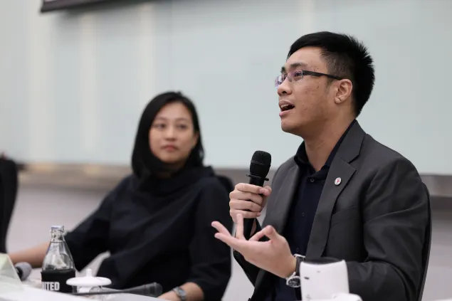 A man speaking into a handheld microphone during a panel discussion, gesturing with one hand. A woman sits beside him, listening attentively. Both are seated at a table with papers and a water bottle in front of them.