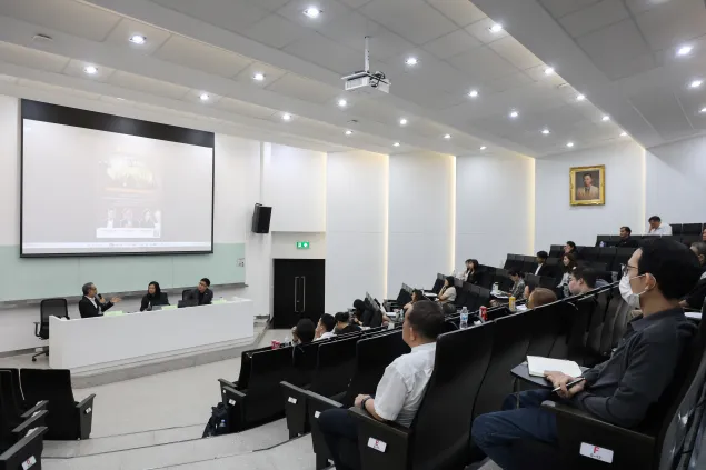 An auditorium filled with attendees seated in tiered rows, watching a panel of speakers at the front. A large screen displays a presentation, and the room is brightly lit with a modern ceiling and projector overhead.
