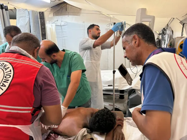 Medical staff and Red Cross volunteers provide emergency care to an injured person lying on a stretcher inside a temporary field hospital, with an IV drip being prepared nearby.