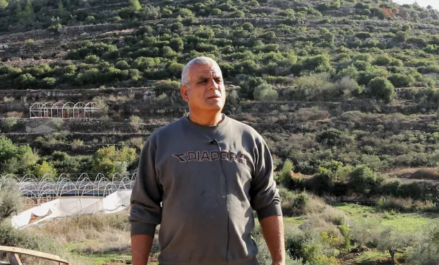 Akram-Radwan está de pie. A sus espaldas, se ven estructuras de invernaderos y una montaña con árboles.