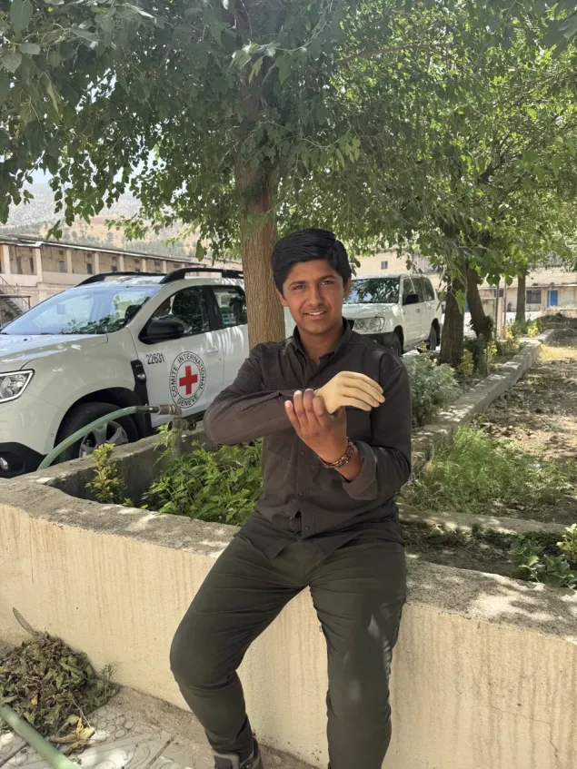 Un joven se encuentra sentado al aire libre, mostrando una prótesis en su mano derecha. Detrás de él se observan vehículos con el emblema de la Cruz Roja.