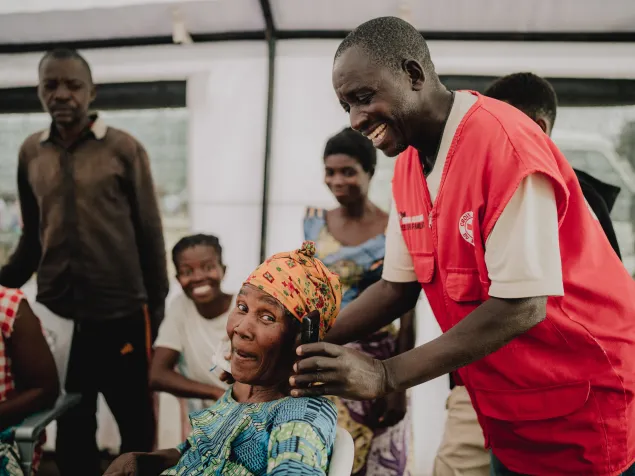  Un trabajador de la Cruz Roja de Burundi ayuda a Maria Chakupwa, refugiada congoleña, a realizar una llamada telefónica dentro de una carpa, mientras otras personas observan alrededor. El trabajador interactúa con ella mientras sostiene el dispositivo, en un esfuerzo por contactar a su sobrina desaparecida.