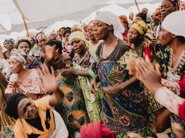 Un grupo de mujeres participa en un servicio religioso en el campamento de Busuma. Varias personas cantan y aplauden, vestidas con prendas coloridas y pañuelos en la cabeza, bajo una estructura cubierta.