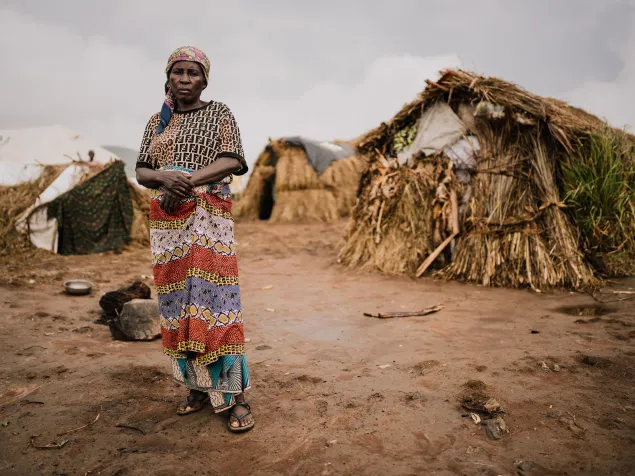 Una mujer se encuentra de pie en un terreno de tierra frente a refugios construidos con materiales naturales en el campamento. Viste una falda colorida, una blusa estampada y un pañuelo en la cabeza. Estelle Tilifoza, originaria de Baraka, viajó con su esposo y sus cinco hijos a través de la ciudad de Rumonge, en el sur de Burundi.