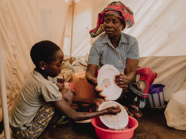Una mujer y una niña lavan platos dentro de una tienda de campaña. La mujer sostiene un plato mientras la niña lo enjuaga en un recipiente con agua. Anastasie Cubwa vive en el campamento de Busuma con su esposo Joseph y sus cuatro hijos.