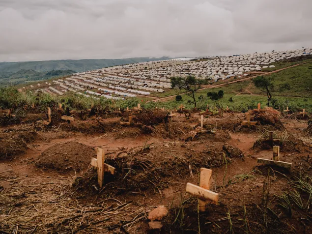 Fotografía en primer plano con tumbas marcadas con cruces de madera en un terreno de tierra. Al fondo, en una colina, se extiende el campamento de refugiados de Busuma con filas de refugios. Un cementerio de personas refugiadas congoleñas fallecidas recientemente se encuentra cerca del campamento de Busuma.