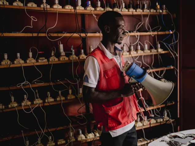 Elyse Bitangimana, trabajador de la Cruz Roja de Burundi, sostiene un megáfono mientras se encuentra en un centro de conectividad del CICR en el centro de tránsito de Cishemere. Al fondo se observan múltiples dispositivos conectados a una red de cables para la carga de teléfonos.