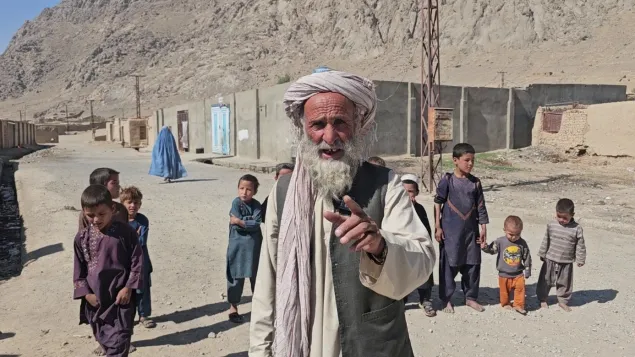 A man wearing a turban gestures toward the camera while standing in a dusty village, with several children and simple buildings set against a mountainous landscape in the background.