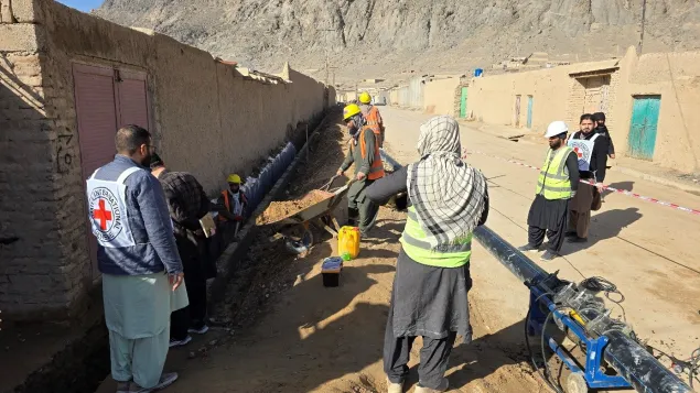People stand along a large water pipe distributing water into yellow jerrycans in a narrow village street, with Red Cross workers assisting and simple buildings lining the scene.