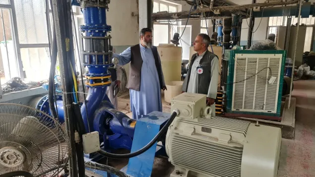 Two men stand inside a technical facility beside large water pump equipment and machinery, discussing operations in a well-lit industrial space.