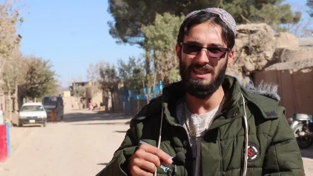A man wearing glasses and a green jacket stands on a sunlit street in a village, speaking while holding an object, with buildings, trees, and a vehicle in the background.
