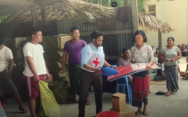 A Red Cross staff member hands a relief item to a woman while others stand nearby outside homes, with bags and supplies gathered for distribution.