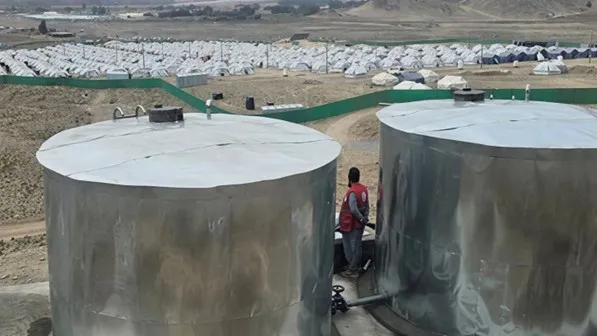 Two large metal water storage tanks stand on a hillside, with a person working between them and a settlement of tents visible in the background.