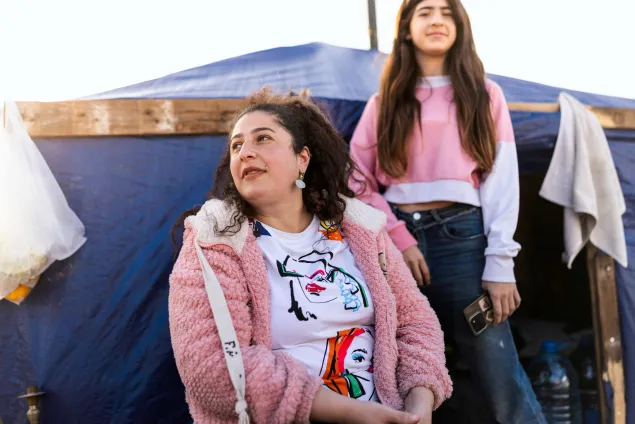 Farah sits outside a makeshift shelter, wearing a pink jacket, while her daughter stands beside her.