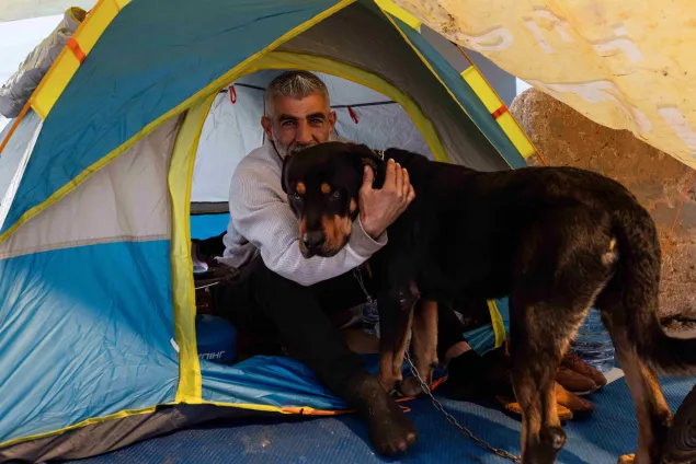 Zaher sits at the entrance of a small tent, holding his dog close, with personal belongings arranged inside the shelter in Lebanon.