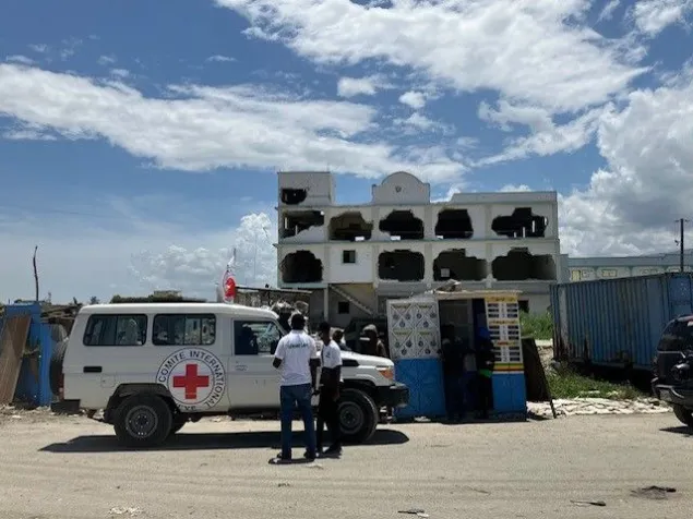 Vehículo del Comité Internacional de la Cruz Roja estacionado frente a una infraestructura de un hospital con daños visibles. Varias personas permanecen junto al vehículo en un entorno urbano afectado por la violencia.