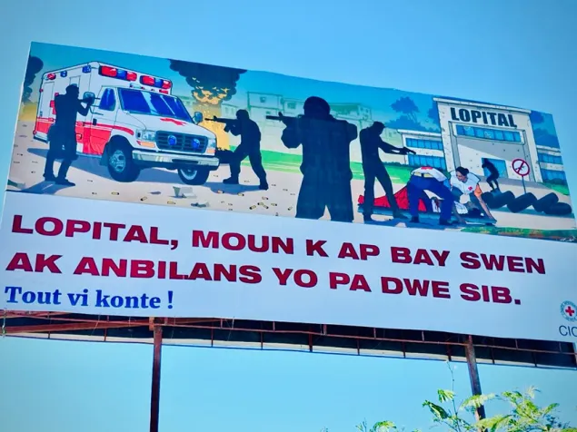 Valla informativa con ilustración sobre la protección de hospitales, personal de salud y ambulancias. El cartel incluye imágenes de una ambulancia, personal sanitario y un centro de salud, junto a un mensaje de sensibilización sobre la importancia de proteger los servicios de salud.