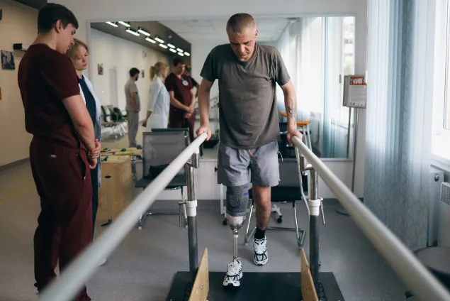 A man uses parallel rails to take steps on a prosthetic leg while physical rehabilitation staff watch on for any support needed.