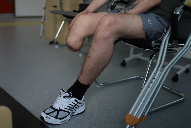 Close up of a man's lower body wearing a sneaker on his left foot, with a healed amputation on his right. Crutches are leaning against a chair in the image.