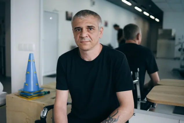A man sitting down in a black t-shirt looks at the camera. 