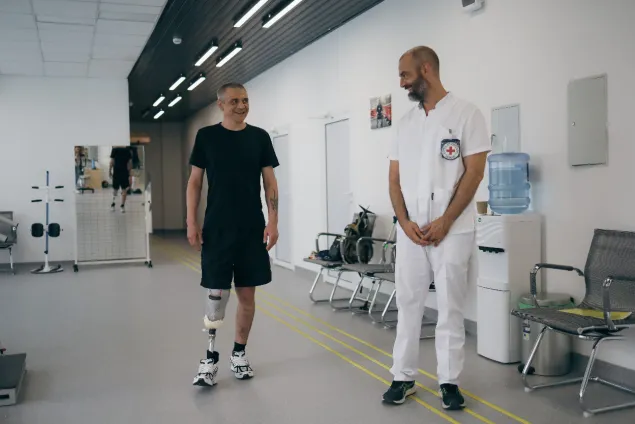 A man with a prosthetic leg is standing and smiling with his prosthetist, who is wearing white scrubs and an ICRC badge.