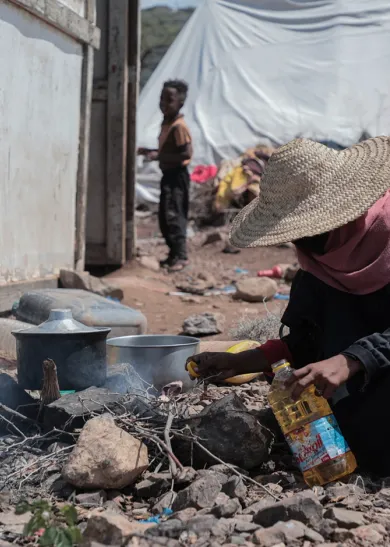 A person cooks over a small outdoor fire among rubble and makeshift shelters.