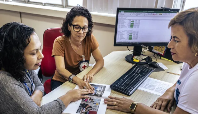 Três mulheres folheiam uma publicação em um escritório; uma delas exibe o logotipo do CICV.