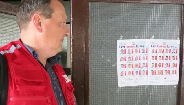 Krnjaca, Sérvia. Cartazes da Cruz Vermelha Sérvia informam os migrantes sobre a possibilidade de buscar os seus familiares. © Cruz Vermelha Sérvia Homem com colete da cruz vermelha olha cartaz com fotos de migrantes.